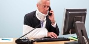 Gewonde zakenman met arm in mitella en nekbrace zit achter zijn computer aan zijn bureau aan de telefoon